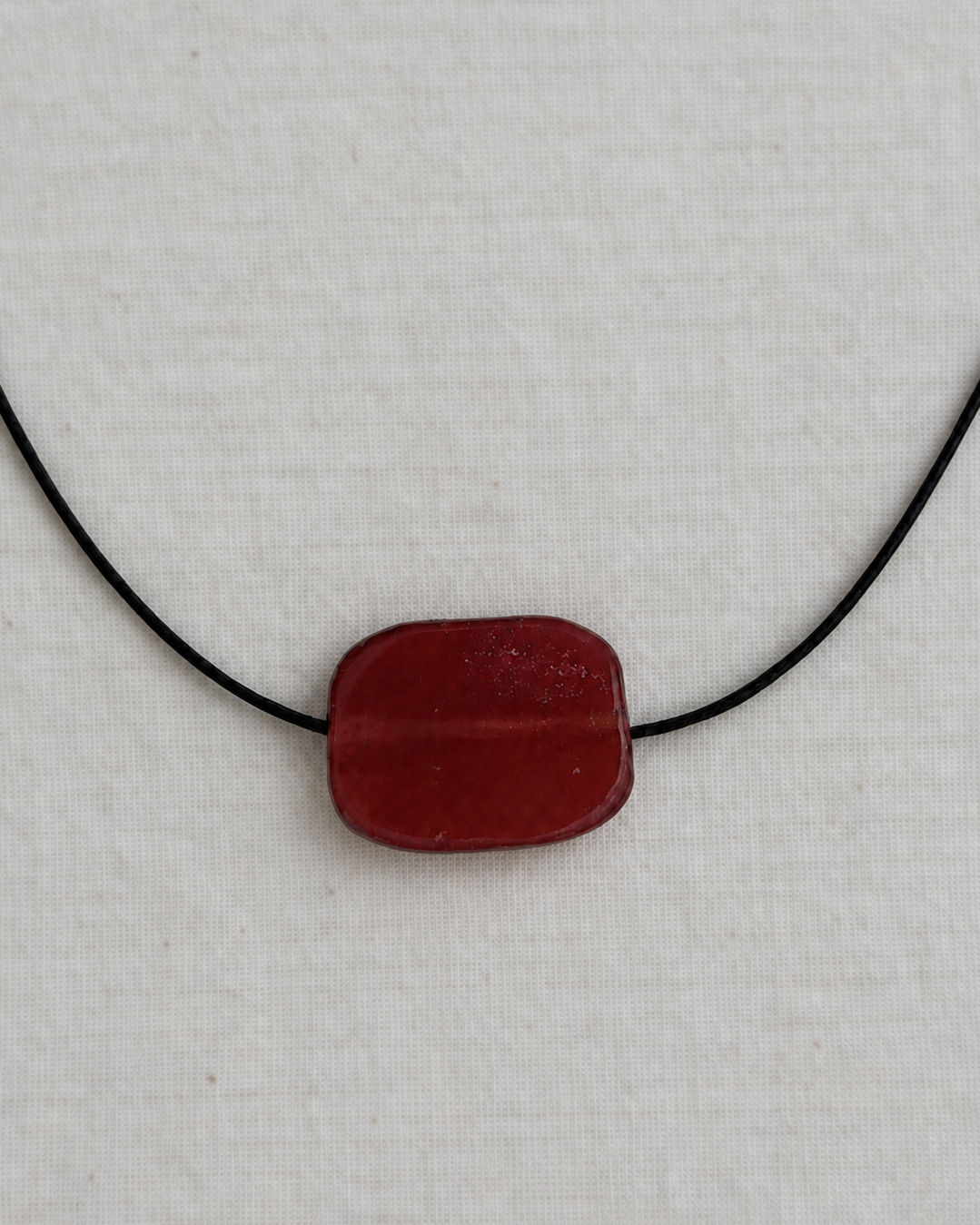 A dark red, rounded rectangular glass pendant on a black cord necklace, displayed against a textured off-white linen background.